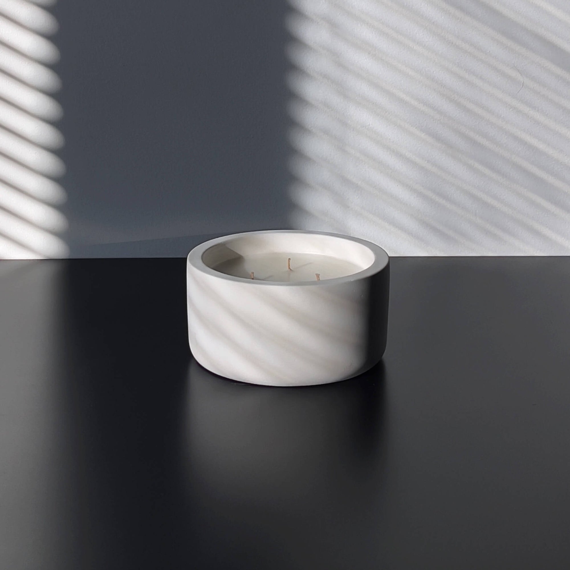 A minimalist, multi-wicked scented candle in a round, smooth concrete vessel. The candle is placed on a dark surface, with soft shadows cast by window blinds against a neutral wall. The clean, simple design emphasizes the modern and tranquil ambiance of the space.