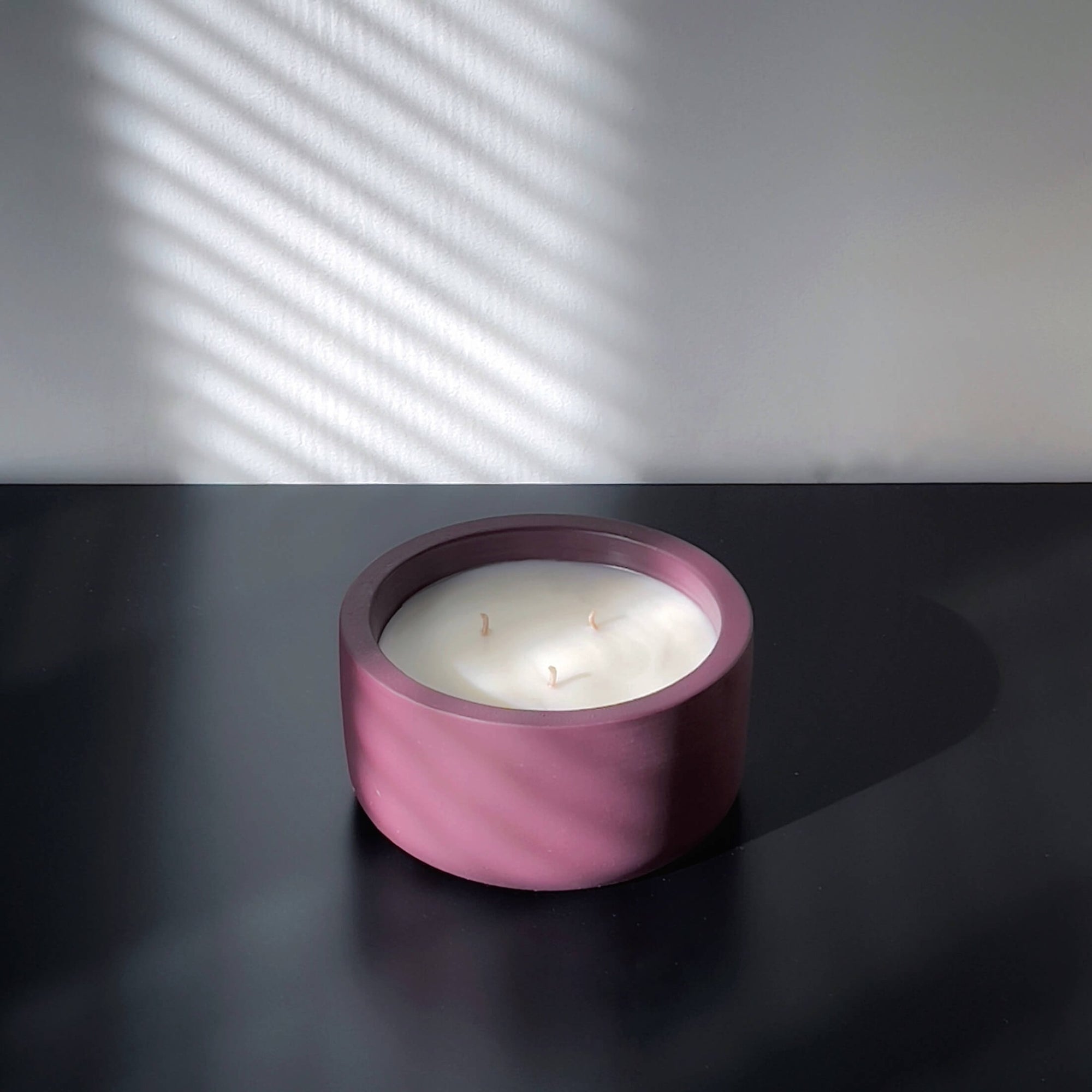 A minimalist, multi-wicked scented candle in a round, deep plum-colored concrete vessel. The candle sits on a dark surface, with soft, diagonal shadows from window blinds cast on the background wall, enhancing the modern, serene aesthetic.