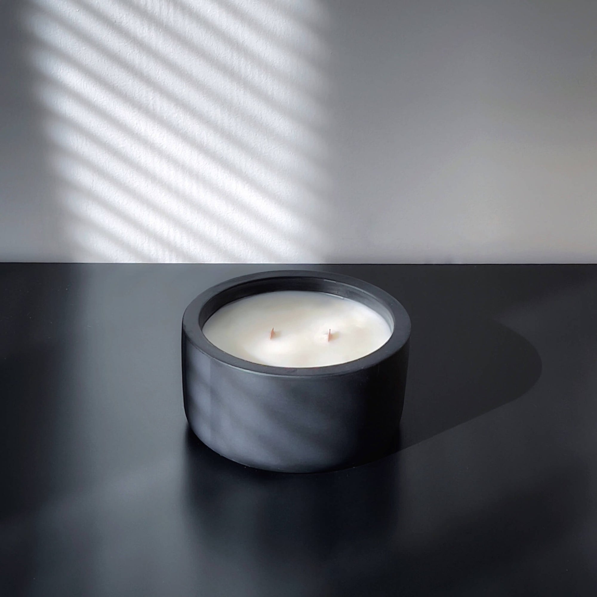 A minimalist, multi-wicked scented candle in a round, matte black concrete vessel. The candle rests on a dark surface, with soft light filtering through window blinds, casting diagonal shadows across the background wall. The sleek, modern design enhances the candle’s luxurious and calming presence.